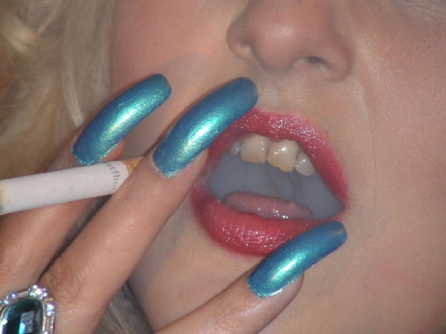 DominatrixChris's face with shiny blue metallic nails, red lipstick, and slightly open mouth, holding a cigarette to her lips. Her teeth are visible. Blonde hair is partially visible.