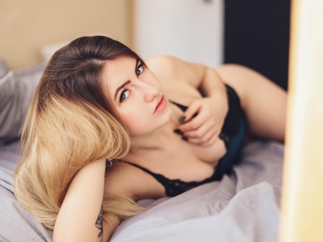ShyNaomi è sdraiata su un letto, indossa biancheria intima di pizzo nero, ha capelli biondi e castani, guarda la telecamera, ha una mano sul petto, mostrando i suoi seni, e un tatuaggio sul braccio..