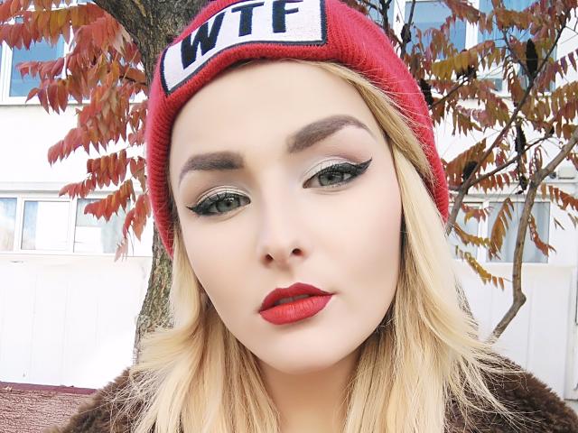 CherrySquirts a la peau pâle, des cheveux blonds, des yeux verts avec du crayon noir, du rouge à lèvres rouge, et porte un bonnet rouge avec l'inscription "WTF". Elle regarde vers le bas avec une expression neutre..
