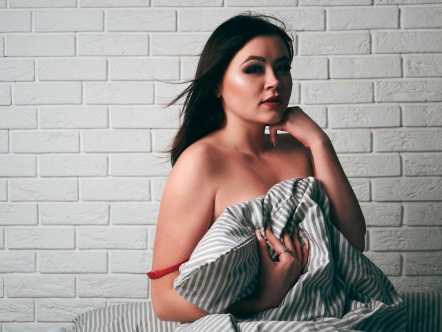 CuttieEyesX, avec ses cheveux foncés, une sangle rouge à l'épaule, tenant une feuille rayée devant ses seins, une main près de sa bouche, un mur de briques blanches en arrière-plan, un léger vent dans ses cheveux, un regard sexy et confiant..