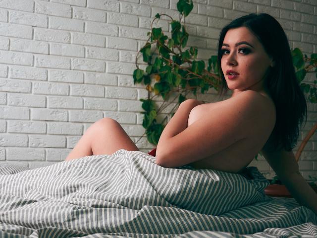 CuttieEyesX, nue, avec les seins couverts par une couverture rayée, est assise sur un lit, les bras croisés, et regarde l'appareil photo. Elle a les cheveux foncés et un mur en briques blanches est visible en arrière-plan..