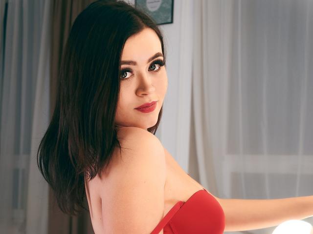CuttieEyesX avec des cheveux bruns foncés mi-longs, un haut rouge sans bretelles, regardant par-dessus son épaule, un sourire doux, du rouge à lèvres rouge, une peau claire, un maquillage subtil, une lumière douce, une légère ombre derrière..