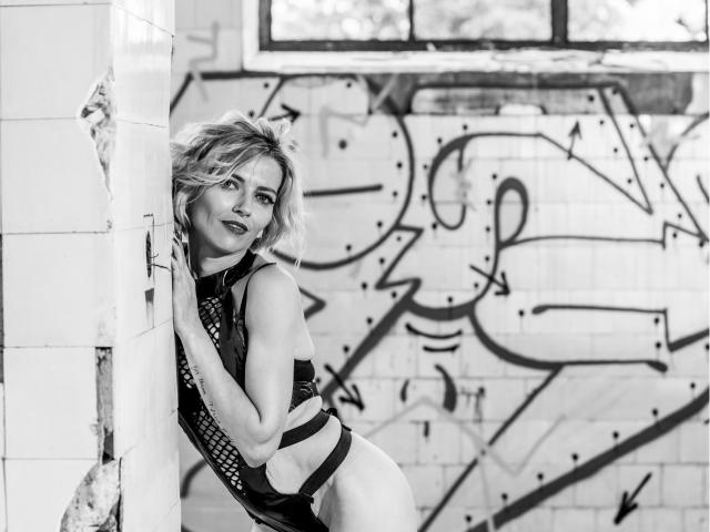 Fotografia in bianco e nero di MoniqDiamond con i capelli biondi, appoggiata a un muro piastrellato, che indossa un outfit di pelle lucida, con spalline sottili e taglio alto, che mette in mostra il sedere e un sorriso sicuro..