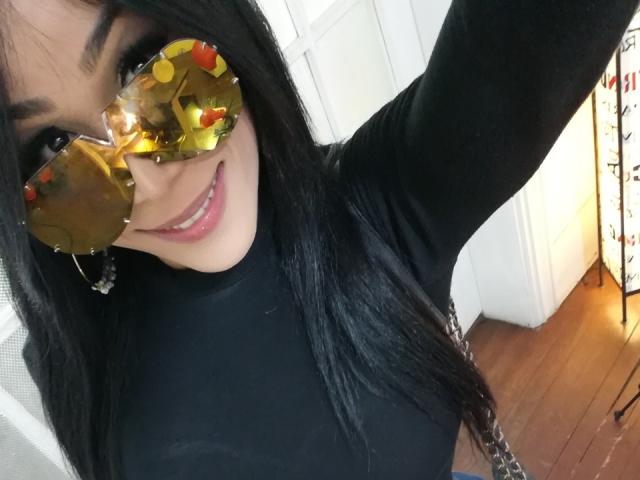LorentsTS with long black hair, wearing yellow reflective sunglasses, smiling, black turtleneck, earrings, raising arm, wooden floor, white door in background. 