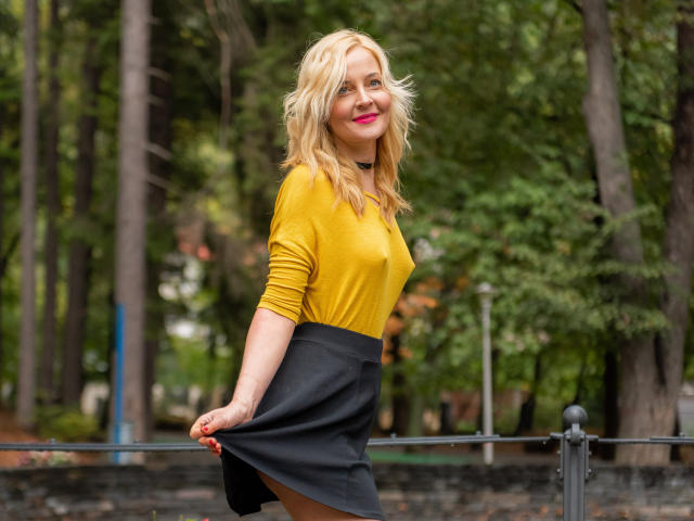 SoleildeNuit con capelli biondi, top giallo, gonna nera e rossetto rosa, sorridente, che tiene la gonna, sfondo di una foresta verde, collana nera, in piedi, smalto per unghie rosso..