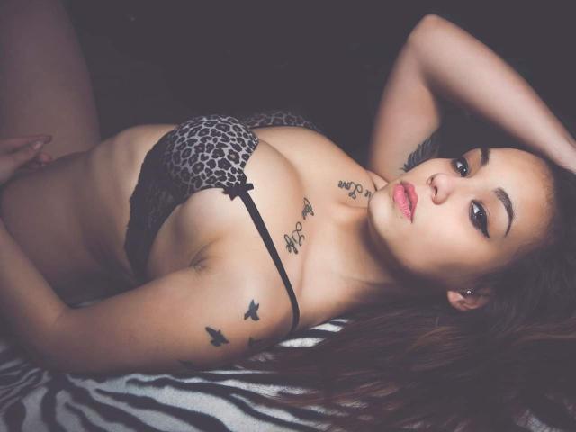 KassiKassou lying on zebra-patterned fabric, wearing a leopard-print bra, exposing her tits, with tattoos on her arm and chest, and a piercing in her ear.