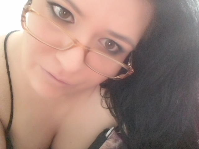 Carolina69 with glasses, brown eyes, and black hair, showing cleavage in a black top, smiling slightly, close-up shot, soft light.