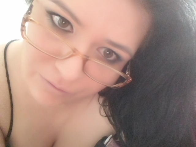Carolina69 with brown glasses, black hair, and light skin, showing her cleavage in a black strap top, smiling slightly at the camera.