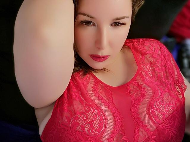 TitanicTits has a fair skin, brown eyes, and red lipstick. She's wearing a red lace top, showcasing her large boobs, with her arm raised. Her expression is sultry.