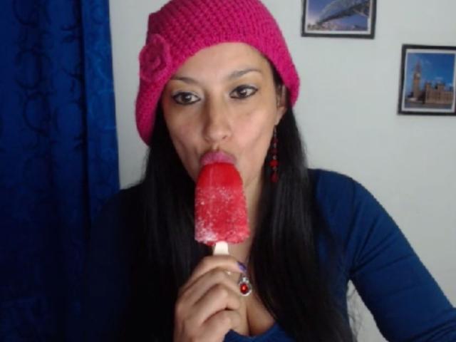WetSweetKim, con lunghi capelli neri, un cappello a maglia rosa e una maglietta blu, che succhia un ghiacciolo rosso e ghiacciato, con gli occhi socchiusi, indossa orecchini rossi e un anello con una pietra rossa..