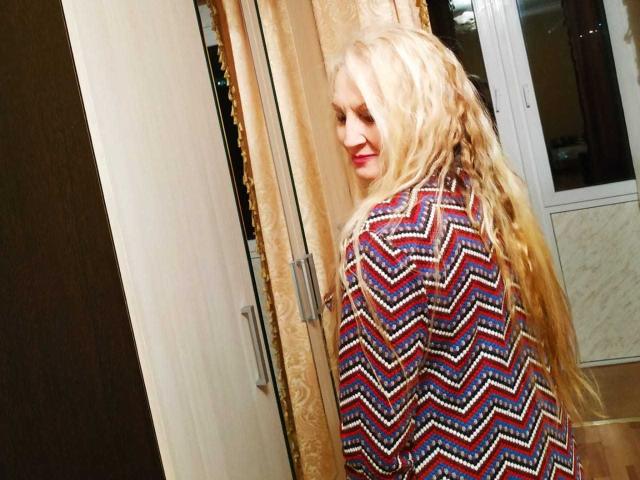 Carly, avec de longues tresses blondes, portant une chemise colorée à motifs en zigzag, souriant par-dessus son épaule, montrant ses fesses dans le reflet d'un miroir..