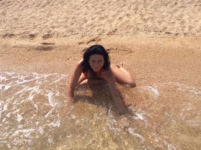 GannaWind with dark curly hair, lying in shallow water, wearing a red bikini, smiling at the camera, with her tits and ass partially above the water.