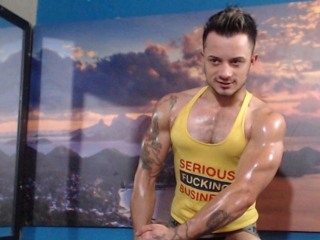 Gespierde Everet met een glanzende, geoliede huid, die een geel tanktop draagt met de tekst "SERIOUS FUCKING BUSINESS", staat met armen over elkaar, met tatoeages op beide armen en kort bruin haar..