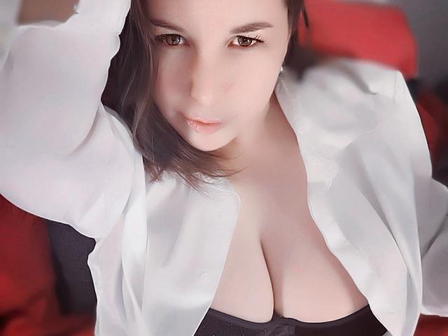 TitanicTits with fair skin, brown eyes, and dark hair, wearing a white shirt open to reveal large, pale tits and a black strap. She looks down with a slightly parted lip.