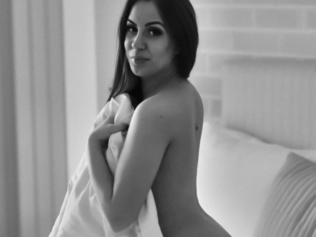 LanieHotXX, topless, with her tits covered by a white sheet, holding it to her chest, looking over her shoulder, in a black and white photo.