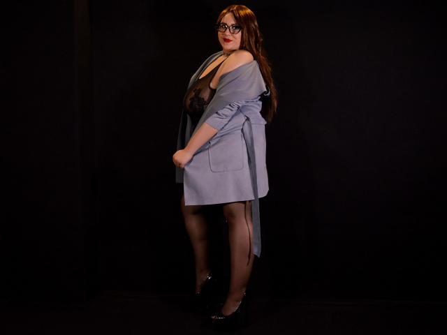 EmmaMelonie, con cabello castaño largo, gafas, un vestido azul que se desliza para revelar lencería de encaje negro, medias negras y tacones negros, de pie frente a un fondo oscuro..