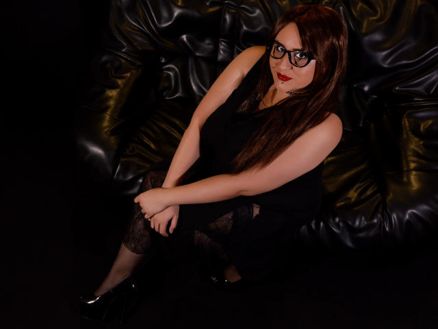 EmmaMelonie, avec des lunettes, du rouge à lèvres rouge, une robe noire, des bas en dentelle et des chaussures noires, assise sur un canapé en cuir noir, regardant vers le haut avec un léger sourire. Ses seins sont petits..