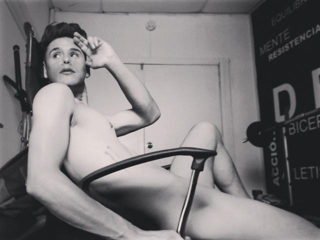 Black-and-white photo of DerrickBigX, naked, sitting in a chair, right hand touching head, left leg raised, small tits, visible ass, short dark hair, looking to the side.