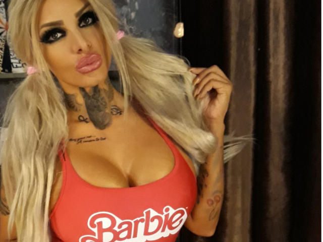BarbaraBae with big tits, blonde pigtails, tattoos, and a red Barbie tank top, pouting with pink lips, holding her hair, in a dark, slightly blurry photo.