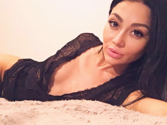 MissTatiana with dark hair, lying on a bed, wearing a black lace top, showing her tits, pink lips, and looking at the camera with a slight smile.
