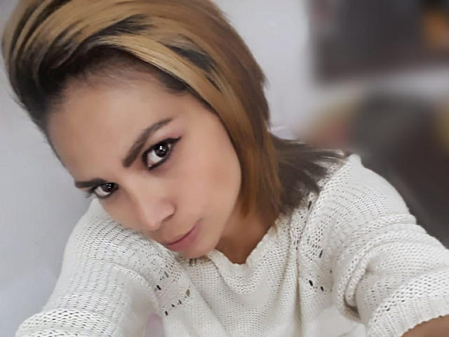 NatashaRomanof with blonde highlights, pouty face, and serious expression, wearing a white, knit, long-sleeve shirt, leaning forward with a slight tilt.