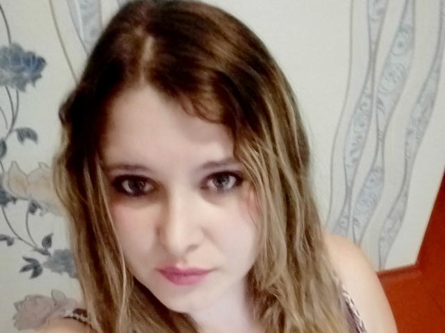 ParisMimi has dirty blonde, wavy hair and light skin with pink lips, staring with brown eyes, wearing a gray lace top, in a selfie with a neutral expression.