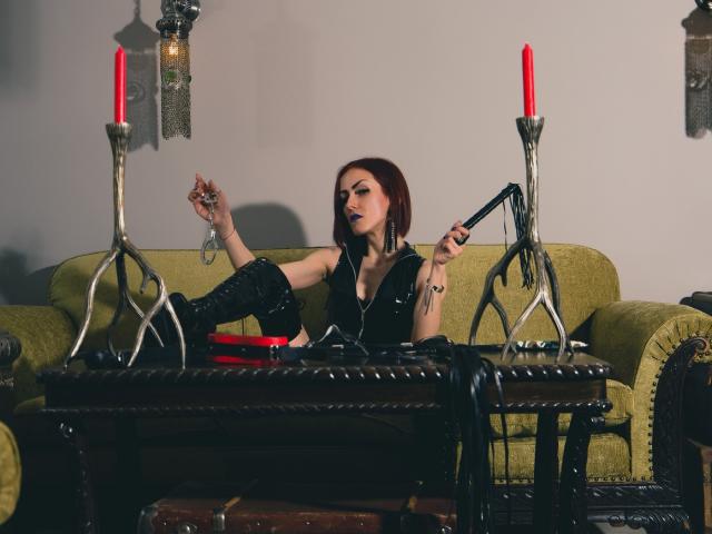 ClaraDomme with red bob, black leather outfit, and boots, holding a whip, sitting on a green couch, arms raised, with two red candles on ornate stands in front.