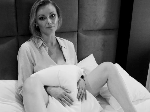 XLovelyVanessa with tits exposed, wearing an open shirt, sitting on a bed with legs spread, holding a pillow over her pussy, smiling, black-and-white photo, looking at camera.