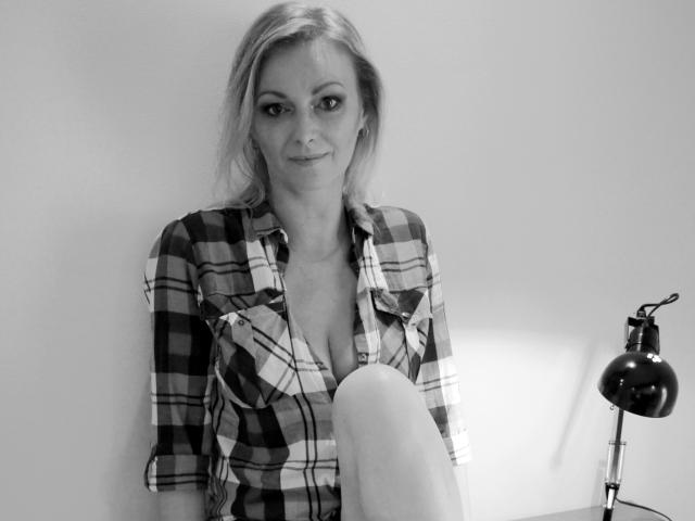 Black-and-white photo of XLovelyVanessa with blonde hair, wearing a plaid shirt, exposing her tits, sitting with one leg up, and a lamp on the right.
