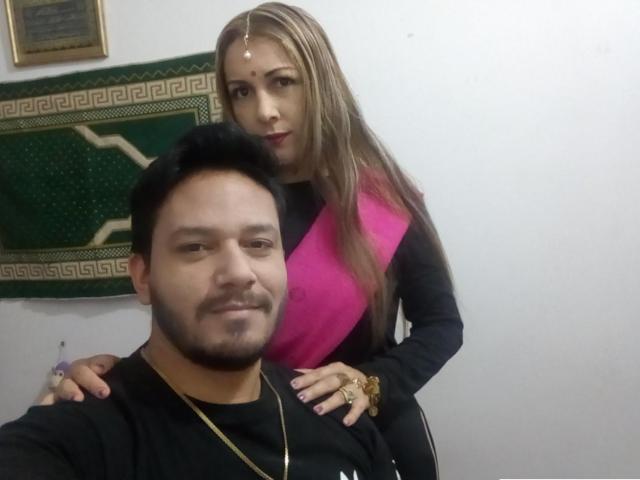 ThorandFreya69, com cabelos castanhos longos, vestindo uma blusa rosa e preta, está em pé atrás de um homem sorrindo, com cabelos pretos, barba e um colar de ouro. A mão dela está apoiada em seu ombro..