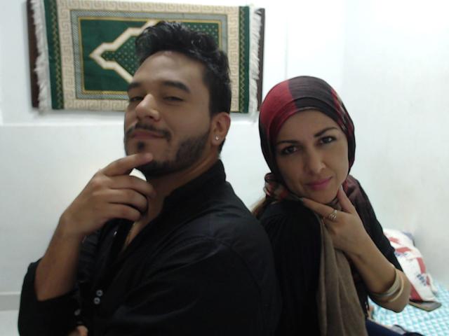 ThorandFreya69, con capelli e barba corti e neri, indossa una camicia nera e ha un dito sulle labbra, sorridendo. Freya69, con un foulard marrone, una camicia nera e un altro foulard marrone, indossa un anello e sorride, con la schiena rivolta verso ThorandFreya.69.