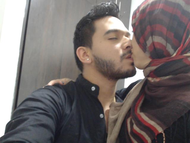 ThorandFreya69 kisses a man with dark hair and a beard, wearing a black shirt, while ThorandFreya69, in a red and brown striped hijab, rests her hand on his shoulder.