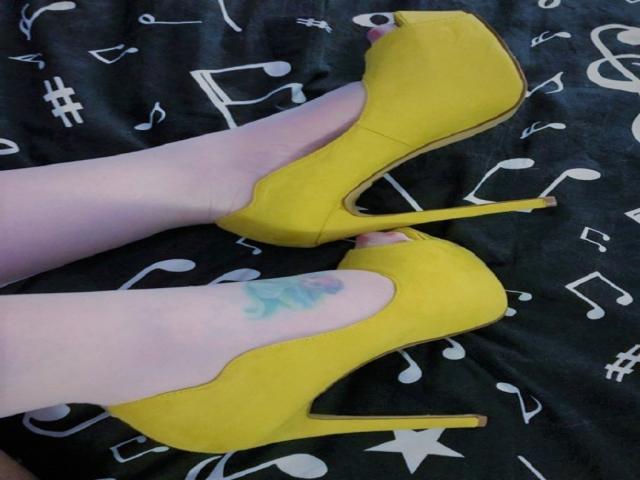 HotKrystall's pale legs in bright yellow stiletto heels, showing a small blue tattoo on the right ankle, with high heels pointed upwards on a dark fabric.