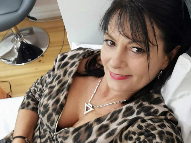 CrystalXBlack, avec ses cheveux noirs, son rouge à lèvres rose et son haut zippé à imprimé léopard qui dévoile sa poitrine, portant un collier en argent, souriant à l'appareil photo..