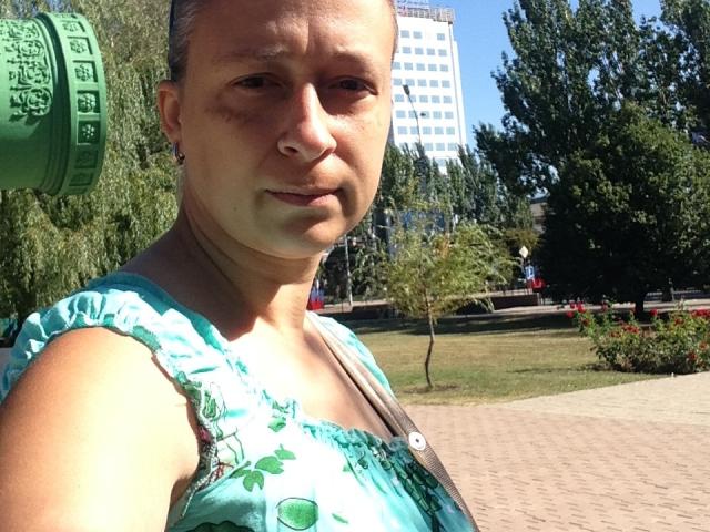 VeneraWings, middle-aged woman with short gray hair, light skin, small nose, wearing teal floral dress, holding green water bottle, standing in sunlit park, looking slightly annoyed.