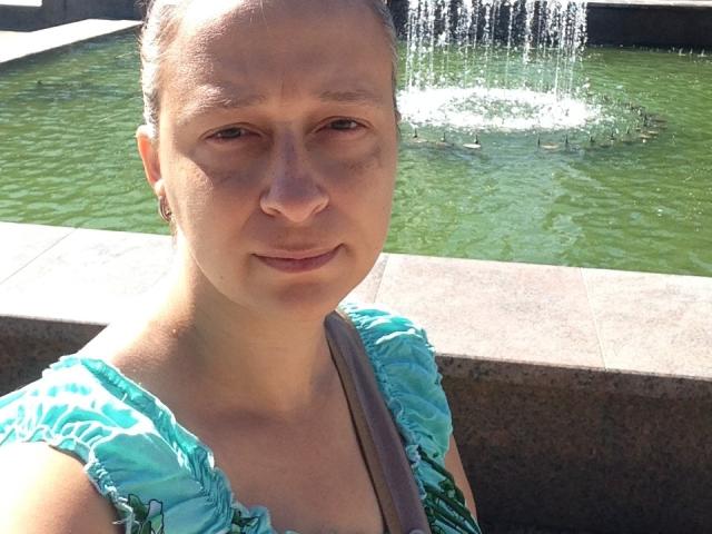 VeneraWings hat helle Haut, braune Augen und kurze, braune Haare. Sie trägt ein türkisfarbenes Kleid mit Rüschen und steht vor einem grünen Wasserspeier. Ihr Hintern ist nicht sichtbar..