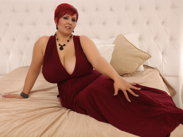 LadyLibely with red hair, large tits, and a deep red dress, sitting on a bed, wearing a black necklace and bracelet, smiling, hand on her dress, tan pillow.