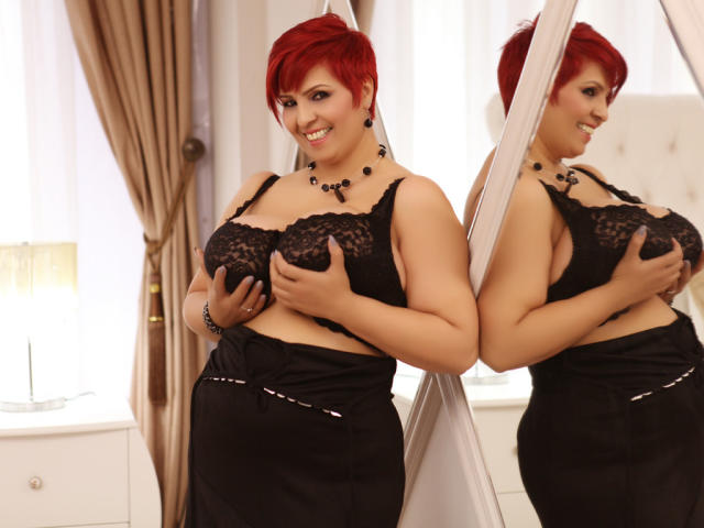 LadyLibely, avec ses cheveux roux, son soutien-gorge en dentelle noire et sa jupe noire, tenant ses seins, souriant en se regardant dans le miroir, portant un collier et des boucles d'oreilles noirs, dans une pièce bien éclairée..