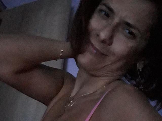 SexyGioconda tiene una ligera sonrisa, cabello castaño, y lleva un sujetador rosa con un collar y pulsera dorados. Su mano está detrás de su cabeza..