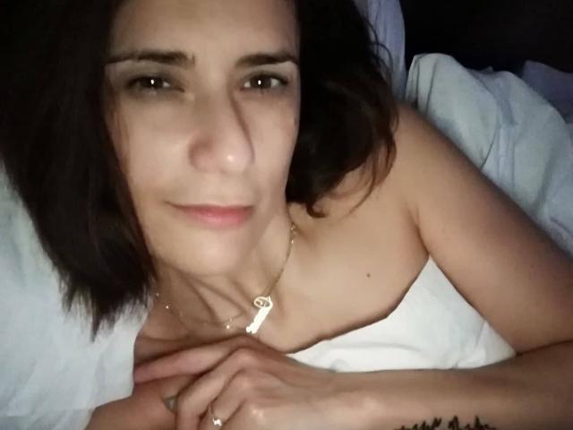 SexyGioconda with short brown hair, light skin, and a subtle smile, lying on white sheets, wearing a strapless top, necklace, and rings, with hands near her chest.