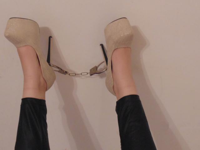 As pernas de BloodyMorticia, vestindo leggings pretas, com saltos altos bege brilhantes e uma alça de corrente dourada, com os pés apoiados em uma parede lisa..