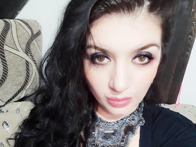 Freyja has pale skin, long black curly hair, purple eyeshadow, brown eyes, pink lipstick, and wears a silver choker with spikes, looking directly at the camera.