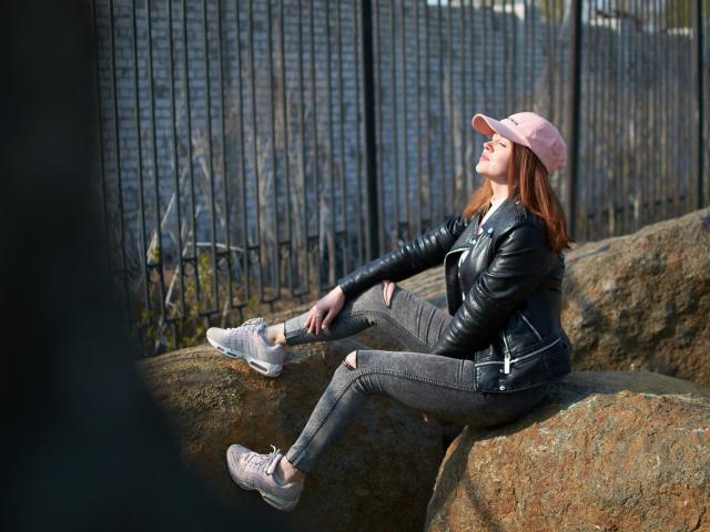 CuttieEyesX con i capelli rossi, un berretto rosa, una giacca di pelle nera, jeans grigi strappati e scarpe da ginnastica rosa, seduta su delle rocce, che si tocca la caviglia, con la luce del sole sul viso..