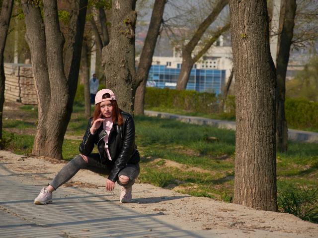 CuttieEyesX, agachada, vistiendo una chaqueta de cuero negra, un sombrero rosa, jeans grises rotos, zapatillas rosas, sosteniendo un teléfono cerca de la boca, en un parque arbolado, con árboles, hierba y un sendero de arena en el fondo..