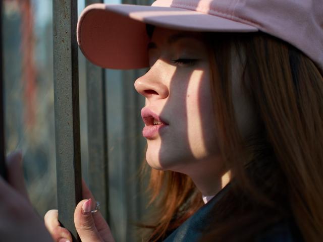 CuttieEyesX con gorro rosa, sosteniendo una barra de metal, sombra en el rostro, labios ligeramente entreabiertos, uñas rosas, cabello castaño, iluminado por el sol, expresión tranquila, luz suave, fotografía realista..