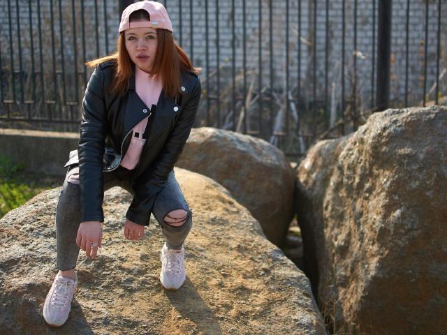 CuttieEyesX mit roten Haaren, einer schwarzen Lederjacke, einem pinken Hemd, zerrissenen Jeans, pinken Turnschuhen und einer pinken Mütze, sitzt auf einem Felsen, im Freien, mit großen Felsen im Hintergrund..