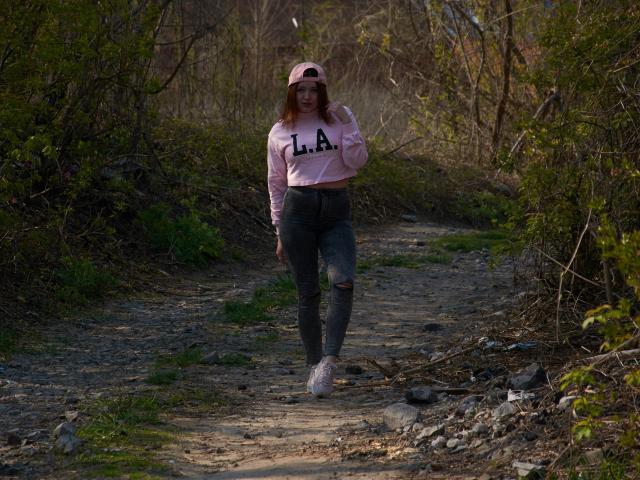 CuttieEyesX walking on a dirt path, wearing a pink "LA" hoodie, gray ripped jeans, pink sneakers, and a pink baseball cap, with a confident, relaxed expression.