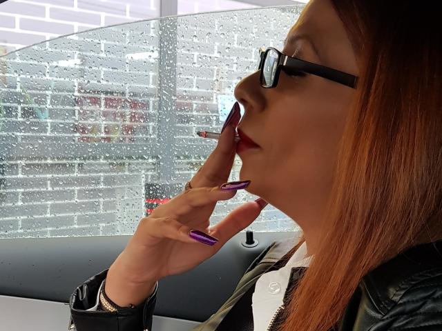 IssaDiamondd, con uñas moradas, gafas negras y lápiz labial rojo, se toca la barbilla, mirando a la derecha, dentro de un coche con lluvia en la ventana, y lleva una chaqueta de cuero negra..