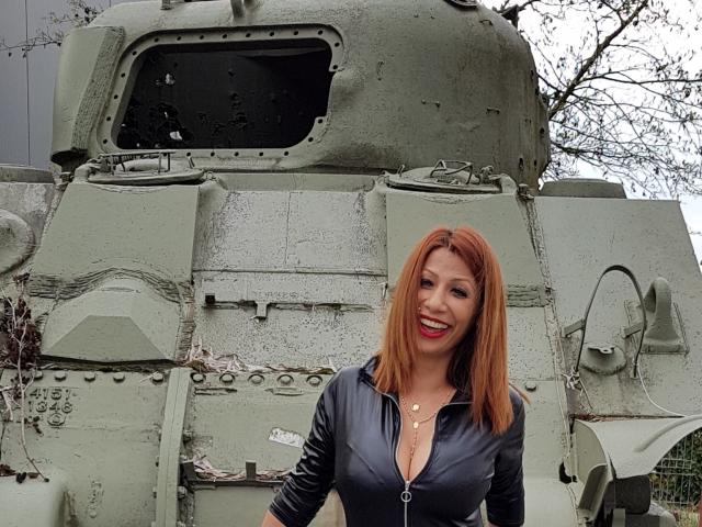 IssaDiamondd mit roten Haaren, lächelnd, trägt ein schwarzes Top mit Reißverschluss und steht vor einem grünen, rostigen, beschädigten Panzer, wobei ihre Brüste leicht durch das Top hindurch sichtbar sind..