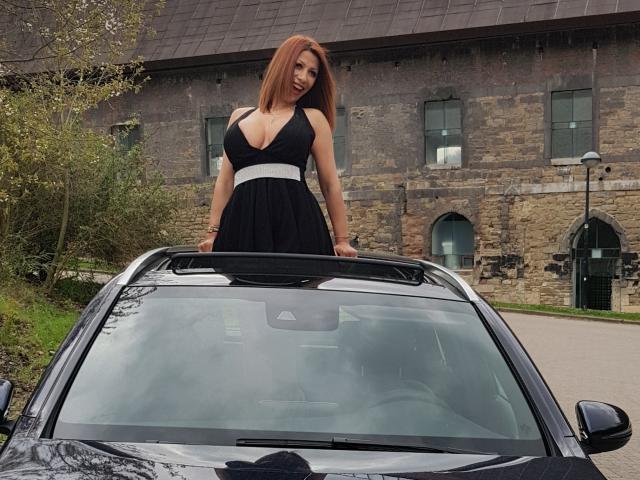 IssaDiamondd mit großen Brüsten, stehend auf einem schwarzen Auto, trägt ein schwarzes Kleid mit einem weißen Bund, vor einem steinernen Gebäude, mit braunen Haaren und rotem Lippenstift..
